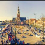 Een bruisend stadsplein met mensenmassa's, marktkraampjes en een centrale kerk. Het beeld heeft een tilt-shift effect, waardoor de scène miniatuur lijkt. Mensen lopen rond onder een heldere hemel, met historische gebouwen op de achtergrond.